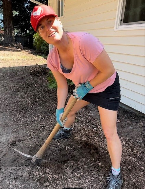 Pema Rocker digs with a pick axe and smiles at the camera in a pink shirt and red baseball cap.